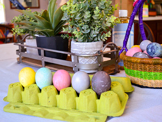 A bunch of finished eggs sitting in an egg carton, with an Easter basket and plant in the background.